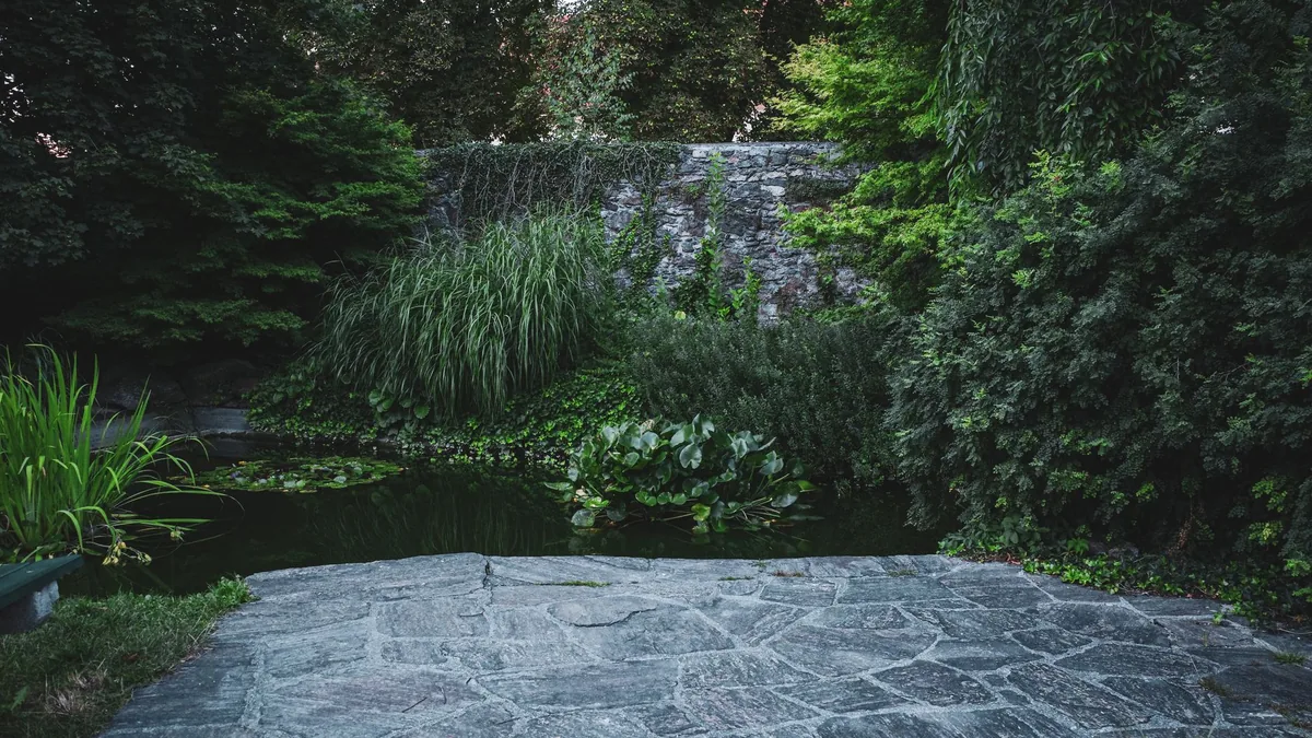 A tranquil garden scene with a stone pathway leading to a pond, surrounded by lush, verdant plants and trees.
